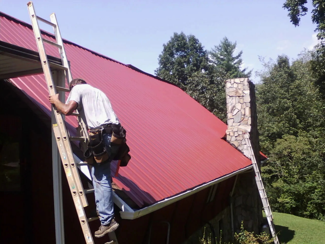 Roofer on a ladder beside a red metal roof and stone chimney — Soffit & Fascia Repair by Roofing Brothers in Estill